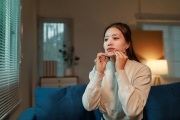 Sitting comfortably on her sofa in the living room, a young Asian woman is applying a moisturizing...