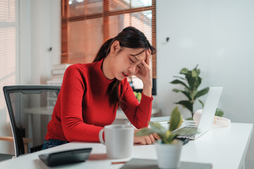 Young woman sitting at her desk, feeling overwhelmed and fatigued while working on a laptop....