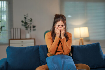 Young woman sitting on the sofa in a dimly lit living room, covering her face with her hands while...