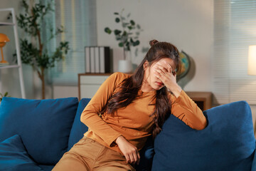Stressed Asian woman sitting alone on a sofa at home, covering her face with her hand while...