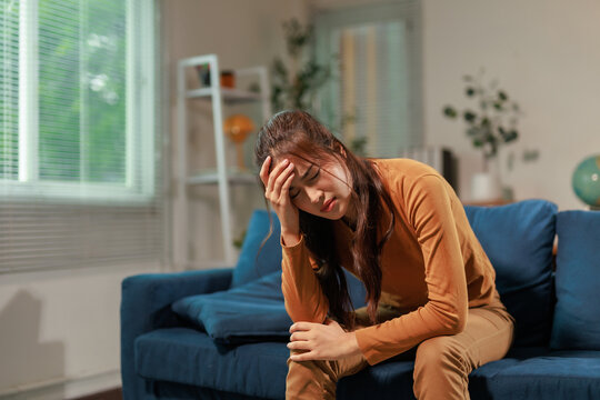 Stressed asian woman having headache and touching her forehead while sitting on the sofa in the living room at home, feeling tired and suffering from chronic migraine