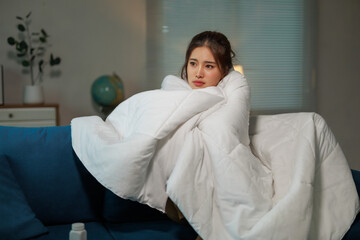 Young Asian woman sitting on a sofa in the living room, wrapped in a cozy white blanket, feeling...
