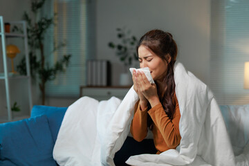 Young woman having a cold, flu or allergy, blowing her nose with a tissue, sitting on a sofa...