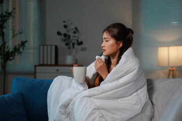 Young asian woman covered in blanket is sitting on the sofa, holding a cup of hot beverage and a...