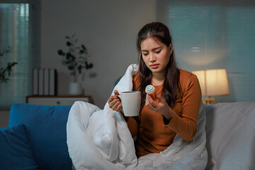 Young Asian woman sitting on a sofa, wrapped in a cozy blanket, sipping tea while reading medicine...
