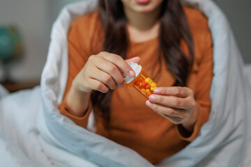 Young woman covered with blanket opening a pill container while sitting in bed at night, taking...