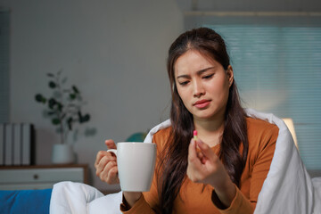 Young Asian woman feeling unwell with the flu, taking medicine while holding a cup of hot tea on...