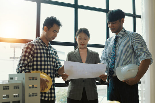 Architects and engineers, wearing hardhats, are reviewing detailed blueprints alongside a scale model of a skyscraper in a bright, modern office filled with natural light