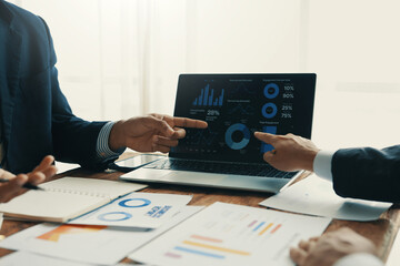 Business people in suits are pointing at a laptop screen displaying financial charts and graphs,...