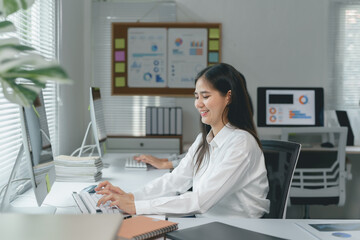 Young Asian businesswoman sitting at desk in modern office, typing on keyboard, working with computer and smiling, with charts and graphs in background