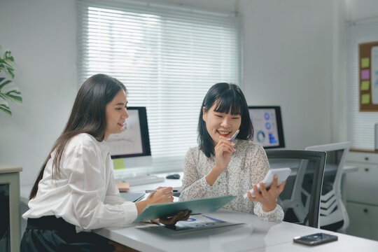 Two joyful businesswomen collaborating and smiling while calculating profits, using a calculator and computer at a bright office desk, showcasing teamwork and professional growth