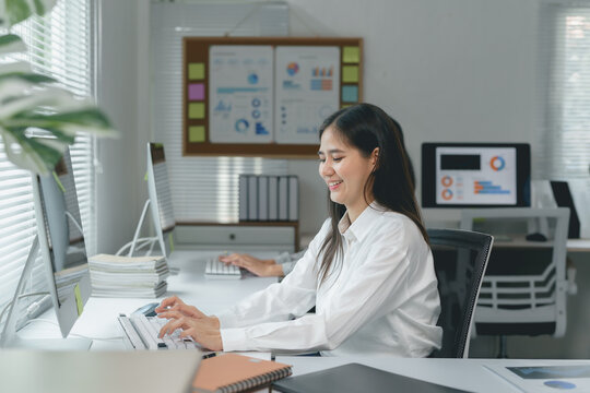 Young Asian businesswoman sitting at desk in modern office, typing on keyboard, working with computer and smiling, with charts and graphs in background