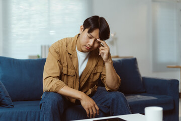Young Asian man sitting on a sofa at home is having a bad headache and feeling stressed, he is...
