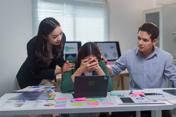 Two colleagues comforting a stressed businesswoman sitting at her desk in the office, showing...