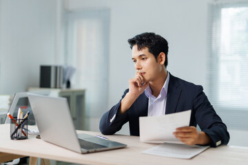 Young businessman holding documents and contemplating his work while seated at a desk with a laptop...