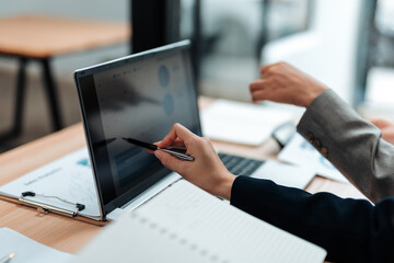 Businesswomen are discussing financial data displayed on a laptop screen, using a pen to point at...