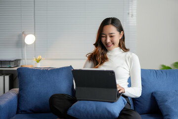 Happy young Asian businesswoman working from home, comfortably sitting on a blue sofa in her living...