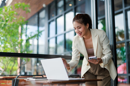 Young asian businesswoman smiling and working using laptop and smartphone in a cafe, she's wearing a white blazer and standing next to a table - Powered by Adobe