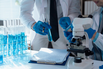 Two scientists wearing lab coats and gloves are conducting research, analyzing blue liquid in a...