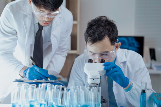 Two asian scientists wearing lab coats and gloves are conducting research in a modern laboratory, one using a microscope and the other taking notes, surrounded by test tubes containing blue liquid