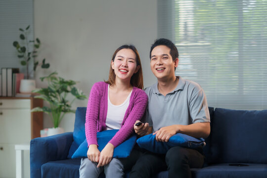 Young asian couple laughing and watching television, enjoying their time together sitting on a comfortable sofa in their modern living room