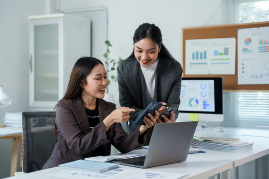 Two happy businesswomen are discussing over a mobile phone and working with laptop and documents on desk at modern office, smiling colleagues discussing new project, business and technology concept