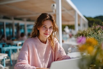 Young woman working on laptop at outdoor cafe near the beach on a sunny day, wearing a pink sweater, surrounded by flowers and modern coastal architecture, enjoying remote work lifestyle