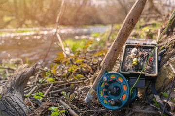 A fly fishing rod and an open fly fishing box lie on the rocks of a mountain river.