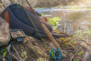 A fly fishing rod and an open fly fishing box lie on the rocks of a mountain river.