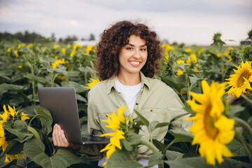 Female agronomist using laptop and inspecting blooming sunflowers in agricultural field