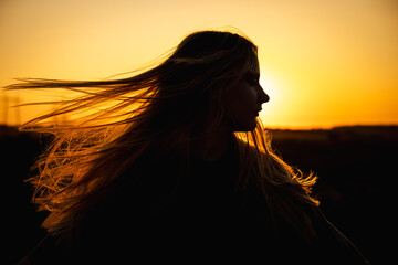 Teen girl in silhouette with flowing hair at sunset, gazing into the distance as warm golden light...