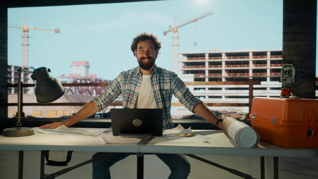 Architect smiling at virtual construction site using laptop and blueprints