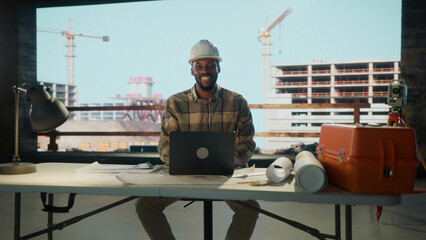 Smiling architect working on laptop at desk with construction site on background