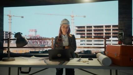 Confident female architect smiling with crossed arms in her office, overseeing construction project with cranes and buildings in background