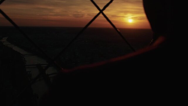 A twilight overview of Paris on sunset as seen from the rooftop through a wire fence, and over the shoulder of a person. Looking on Paris skyline in the evening from a lookout point.