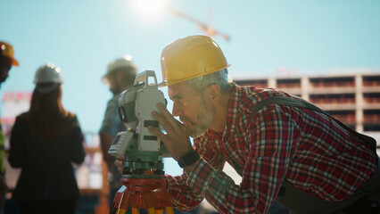 Land surveyor using theodolite, measuring construction site progress with team
