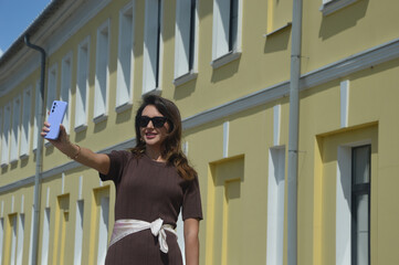 Stylish Caucasian young woman in sunglasses and brown dress taking photo of herself on smartphone...