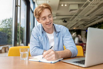 Ginger young man sitting at the table in the cafe and making notes