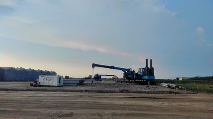 A construction site with heavy equipment used for installing building pillars, surrounded by stacked materials and open land under a wide, blue evening sky.