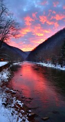 Winter river reflecting a fiery sunset. Mountains border the water, capped with snow. Bare trees line the banks, the sky ablaze with vibrant pink and orange hues
