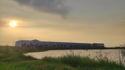 A block of warehouses stands near a quiet pond, bathed in golden hour light. Tall grass sways in the foreground, adding a natural contrast to the industrial setting.