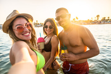 Happy friends taking selfie while enjoying cocktails in the sea at sunset