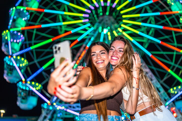 Two happy friends taking selfie in front of ferris wheel at night