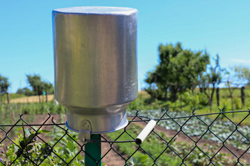 A large metal milk churn hangs on a chain-link fence against a blurred rural landscape with trees...