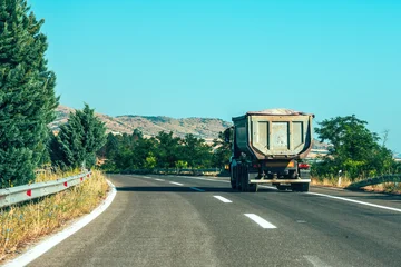 Fototapeten Türkis Dump truck driving on a curvy rural road through dry countryside landscape with trees and distant hills under clear blue sky.  © Bits and Splits