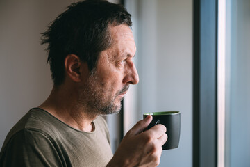 Drinking hot tea or coffee while looking out and thinking, portrait of mature adult male at home