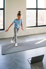 Child practicing balance on a mat while following instructions from a laptop in a bright room