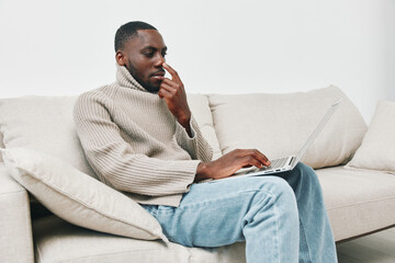 Young Black man in a cozy beige sweater sitting on a sofa, thoughtfully using a laptop in a modern,...