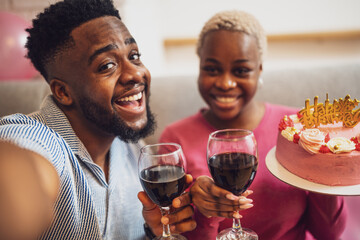 Happy young couple celebrating woman's birthday at home. Woman is holding cake and they are toasting with wine.