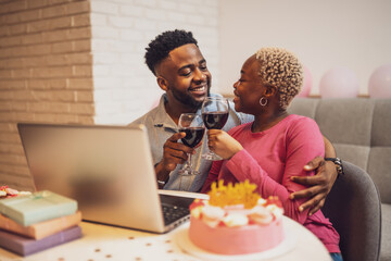 Happy young couple celebrating woman's birthday at home. They are toasting with wine and having video call with family.